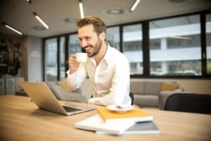 Smiling man in office drinking coffee and reading renewable energy email newsletters on his laptop.